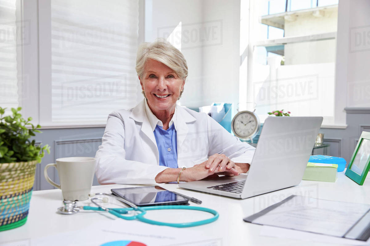 Female Doctor Sitting At Desk Working At Laptop In Office Stock Photo